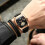 Relógio masculino com pulseira de couro marrom e preta, indicador de dia e data, resistente à água e com fase da lua. Design quadrado com 5